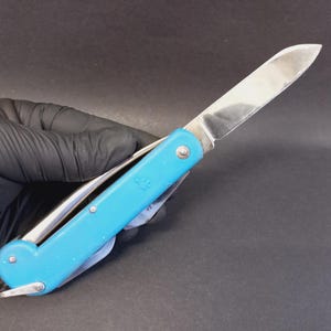 May include: A folding pocket knife with a bright blue handle and a single stainless steel blade extended. The handle features a small embossed logo of a hand and a metal loop at the base. The knife is held in a black gloved hand.