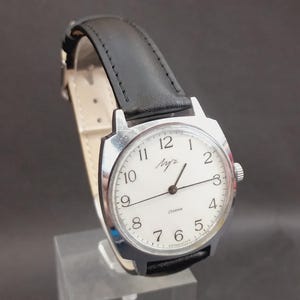 May include: A wristwatch with a silver-toned metal case and a white dial featuring black Arabic numerals. The watch has thin black hands and a black leather strap with visible stitching. The dial displays the brand name in script text.