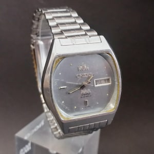May include: A silver-toned Orient wristwatch featuring a grey dial, a metal link bracelet, and a day-date window at the three o'clock position. The watch face displays the Orient logo, the text 'AAA Crystal', and '21 Jewels' with silver hour markers.