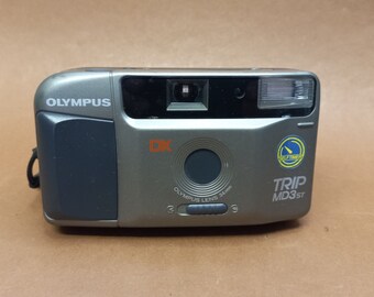 May include: A silver Olympus Trip MD3ST DX point-and-shoot film camera with a black lens and a self-timer button. The camera has a built-in flash and a black rubber grip.