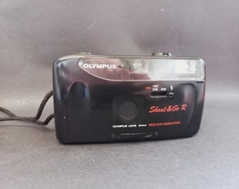 May include: A black Olympus Shoot & Go R 35mm film camera featuring a built-in flash, a 34mm lens, and red-eye reduction technology. The front of the camera displays the brand name, model name, ISO settings, and a black wrist strap.