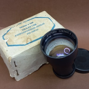 May include: A black film projector lens with a silver lens and the text "N7675954 СДЕЛАНО В СССР" printed on the side. The lens is in a white box with the text "СДЕЛАНО В СССР ОБЪЕКТИВ КИНОПРОЕКЦИОННЫЙ РО-109-1а" printed on the top.