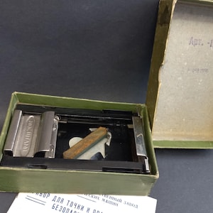 Pode incluir: Uma caixa verde-oliva aberta contendo um afiador de barbear de metal vintage. A caixa é retangular com uma tampa articulada, e o interior contém o afiador, uma pedra de amolar e um suporte de metal. A tampa tem texto em língua estrangeira.