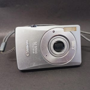 May include: A silver Canon Digital IXUS 75 compact camera featuring a 3x optical zoom lens, a built-in flash, and a 7.1 megapixel sensor. The device includes a grey wrist strap attached to the side and displays the brand name and model text.