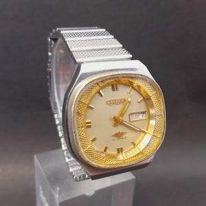 May include: A silver-tone Citizen automatic wristwatch featuring a cream-colored dial with gold-tone hour markers and hands. The watch has a textured gold-tone bezel, a day-date window at the three o'clock position, and a flexible silver-tone metal link band.