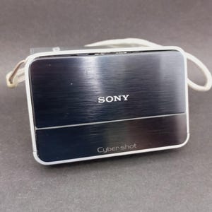 Puede incluir: Una cámara digital Sony Cyber-shot rectangular con un acabado metálico oscuro cepillado y un borde blanco. El dispositivo cuenta con una cubierta frontal deslizante con el logotipo de Sony centrado y el logotipo de Cyber-shot en la parte inferior, junto a una correa blanca.