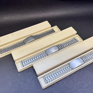 May include: Four silver-toned watch bands are displayed in off-white rectangular boxes. The watch bands have a linked design with a rectangular clasp. The boxes are arranged diagonally, with the bands visible.