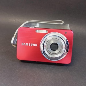 Puede incluir: Una cámara digital Samsung roja con acabado de metal cepillado, barril de lente plateado y flash integrado. La cámara presenta un logotipo blanco de Samsung en la parte frontal e incluye una correa de muñeca gris claro en el lateral.