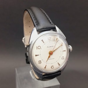 Peut inclure: Une montre-bracelet vintage avec un boîtier argenté et un cadran crème. La montre présente des chiffres noirs, des aiguilles dorées et un bracelet en cuir noir. Le cadran de la montre porte le mot "AAMAZ".