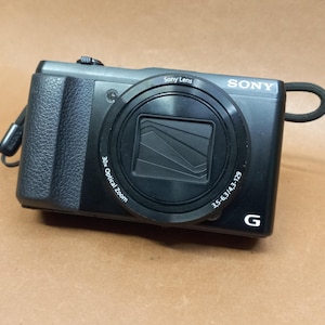 May include: Black Sony digital camera with a prominent lens and a textured grip. The camera has a 30x optical zoom and a wrist strap. The Sony logo is visible on the top right corner. The camera is on a brown surface.