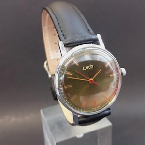 May include: A vintage wristwatch with a black leather band and a silver-colored case. The watch face is a dark olive green with gold-colored hands and the word "Luch" printed in gold. The watch is displayed on a clear stand.