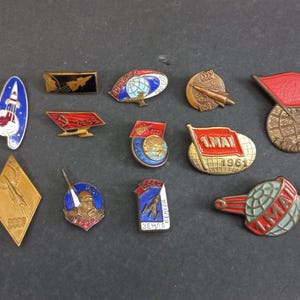 Könnte beinhalten: Eine Sammlung von Vintage-Anstecknadeln aus der Sowjetzeit mit verschiedenen Designs. Die Anstecknadeln zeigen Raketen, Flaggen, Globen und den Text "1.MAI 1961" und "CCCP". Die Anstecknadeln sind in verschiedenen Formen und Farben, darunter Rot, Blau, Gold und Weiß.