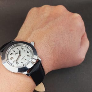 May include: A silver-toned wristwatch with a white face and black leather band. The watch face features black numerals and hands, with the brand name in a stylized font. The watch is worn on a wrist against a dark background.