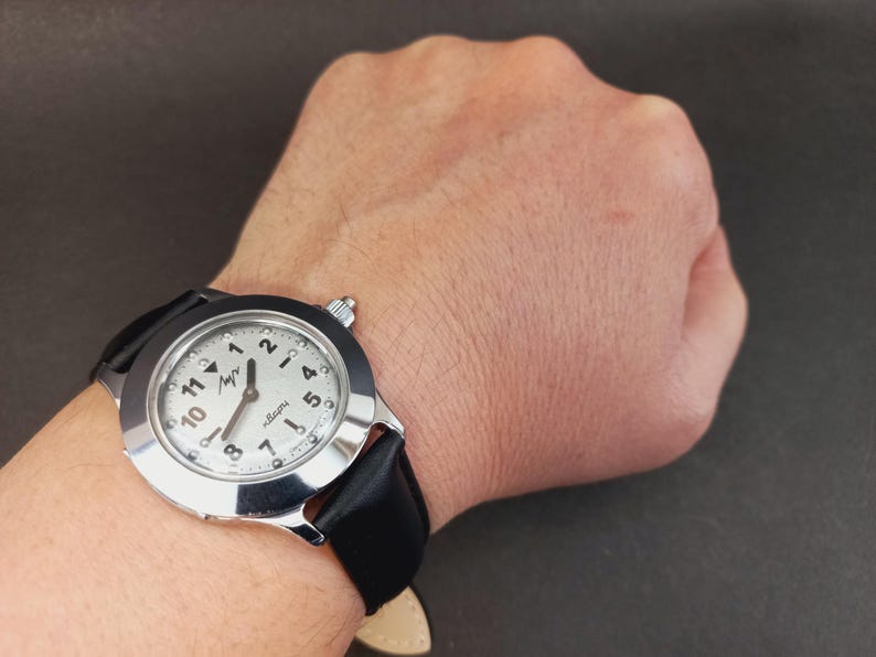 May include: A silver-toned wristwatch with a white face and black leather band. The watch face features black numerals and hands, with the brand name in a stylized font. The watch is worn on a wrist against a dark background.