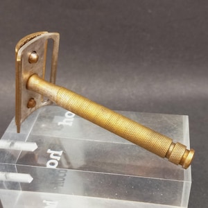 Könnte beinhalten: Ein Vintage-Sicherheitsrasierer mit einem strukturierten, goldfarbenen Metallgriff mit kreuzschraffiertem Muster. Der Rasierkopf besteht aus silberfarbenem Metall und verfügt über zwei kleine kreisförmige Nieten auf der Platte und einen Schutzbügel.