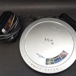 Könnte beinhalten: Silberner Sony CD Walkman MP3-Player mit schwarzem Netzteil und Kabel. Auf dem Player steht "CD WALKMAN" und "NE720". Außerdem gibt es einen Aufkleber mit der Aufschrift "DIGITAL AMP".