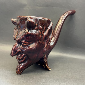 May include: A brown ceramic pipe shaped like a devil's head. The pipe has a long, curved stem and a detailed face with horns.