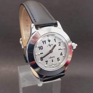 May include: A silver-toned wristwatch with a white face and black numerals. The watch has a black leather band and a small dial on the side. The watch face has the word "Лучь" and "кварц" printed on it.