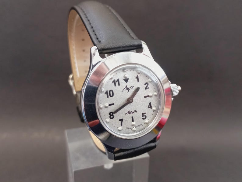 May include: A silver-toned wristwatch with a white face and black numerals. The watch has a black leather band and a small dial on the side. The watch face has the word "Лучь" and "кварц" printed on it.