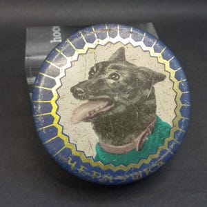 Op de afbeelding: Ronde, vintage metalen blikken doos met een geschilderde illustratie van een zwarte hond met een groene trui en een bruine kraag. De doos heeft een blauw-gouden decoratieve rand en Cyrillisch schrift.