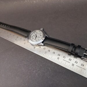 May include: A silver-toned wristwatch with a black leather band. The watch face has black numerals and hands against a light background. The watch is positioned next to a metal ruler, showing measurements in inches.