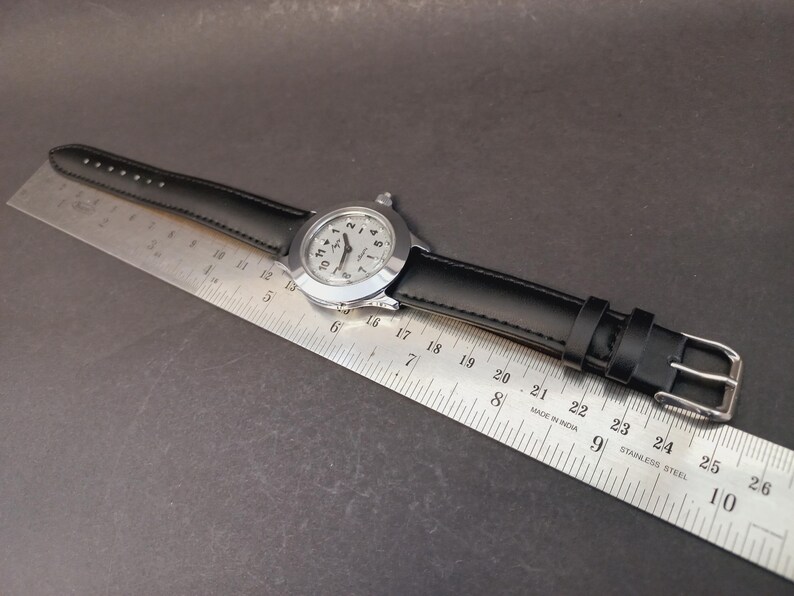 May include: A silver-toned wristwatch with a black leather band. The watch face has black numerals and hands against a light background. The watch is positioned next to a metal ruler, showing measurements in inches.