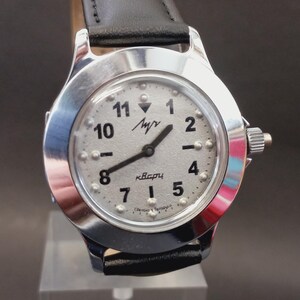 May include: A silver-toned wristwatch with a light-colored face and black hands. The watch face features black numerals and raised dots, with the text "Луч" and "кварц". The watch has a black leather band.