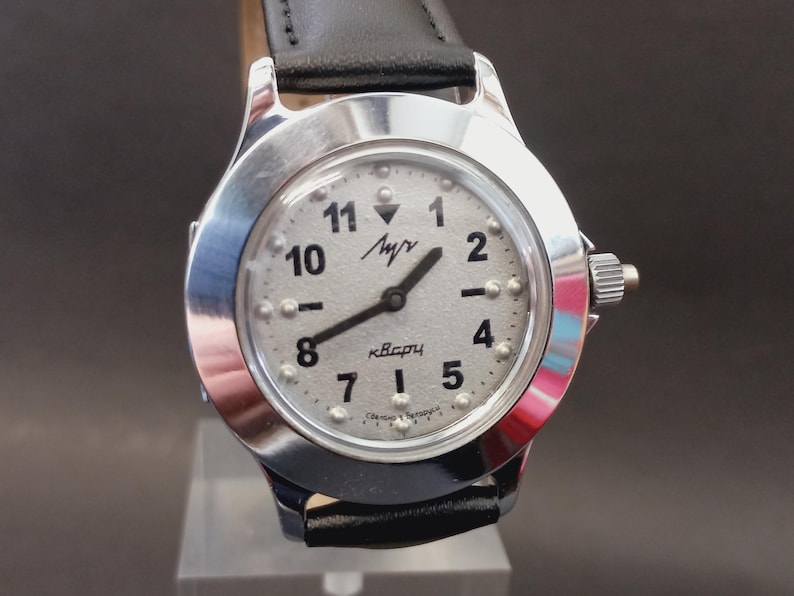 May include: A silver-toned wristwatch with a light-colored face and black hands. The watch face features black numerals and raised dots, with the text "Луч" and "кварц". The watch has a black leather band.