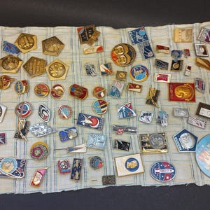 May include: A collection of vintage pins and badges, featuring various shapes and designs. The pins display a range of colors, including gold, red, blue, and silver. Some pins have text, and the collection is arranged on a striped fabric.