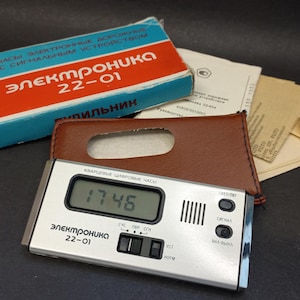 Puede incluir: Un reloj digital vintage con una esfera plateada, que muestra "17:46". El reloj está etiquetado como "Elektronika 22-01" y viene con una funda de cuero marrón y embalaje original. La caja es azul y roja.