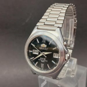 Puede incluir: Un reloj de pulsera Orient plateado con esfera negra, que cuenta con una correa de eslabones de acero inoxidable, marcadores de hora plateados, números romanos en las doce y las seis, ventana de día-fecha y el texto Orient, Water Resist y 50M.