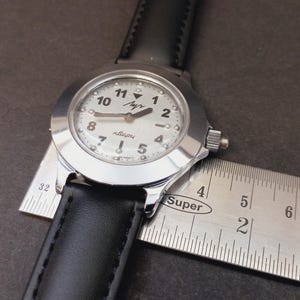 May include: A silver-toned wristwatch with a white face and black leather band. The watch face displays black numerals and hands. A ruler is placed next to the watch to measure its size.
