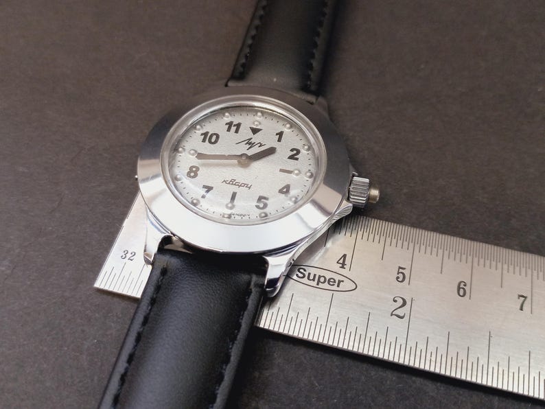 May include: A silver-toned wristwatch with a white face and black leather band. The watch face displays black numerals and hands. A ruler is placed next to the watch to measure its size.