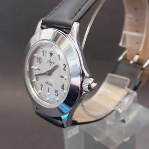 May include: A silver-toned wristwatch with a white face, black numerals, and a black leather band. The watch face displays the word "luch" and the word "quartz". The watch is displayed on a clear stand.