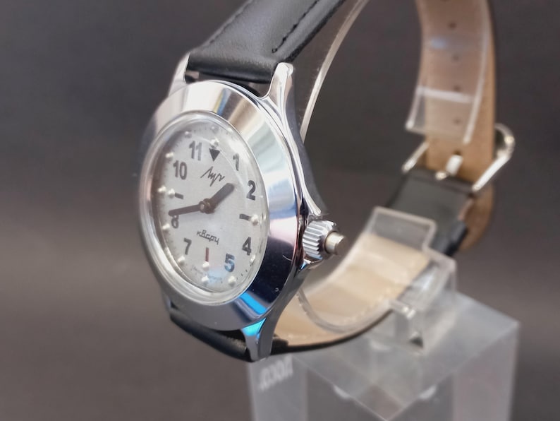 May include: A silver-toned wristwatch with a white face, black numerals, and a black leather band. The watch face displays the word "luch" and the word "quartz". The watch is displayed on a clear stand.