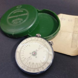 May include: A vintage circular slide rule with a cream face and black markings, in a green plastic case. The case is open, showing the slide rule and a piece of paper with handwritten notes. The slide rule has a metal frame.