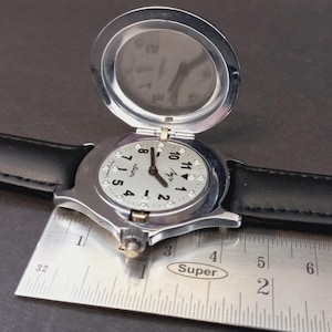 May include: A silver-toned wristwatch with a black leather band. The watch face is light-colored with black numerals and small crystals. A hinged cover is open, revealing the watch face. A metal ruler is placed next to the watch.
