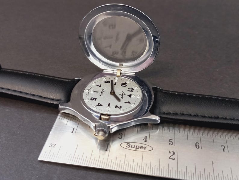May include: A silver-toned wristwatch with a black leather band. The watch face is light-colored with black numerals and small crystals. A hinged cover is open, revealing the watch face. A metal ruler is placed next to the watch.
