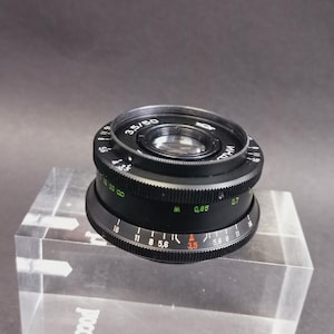 May include: A black camera lens with white and green markings, including the text "3.5/50". The lens is set on a clear acrylic block against a dark background. The lens has a series of numbered rings.