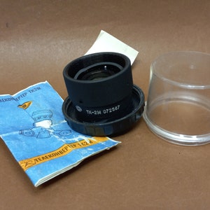 Puede incluir: Un objetivo de teleconvertidor negro con el texto "TK-2M 072387" impreso en el lateral. El objetivo está en una caja de plástico transparente.