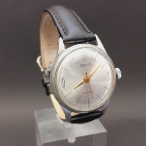 Peut inclure: Une montre-bracelet vintage avec un cadran argenté, des aiguilles dorées et une trotteuse rouge. La montre porte le mot "Восток" sur le cadran et un bracelet en cuir noir. La montre est exposée sur un support transparent.