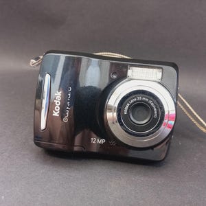 May include: A black Kodak Easyshare digital camera with a 12 MP sensor and a silver-rimmed lens labelled Kodak Lens 35 mm (Equivalent). The compact device features a built-in flash, a silver accent strip on the left side, and a thin wrist strap.