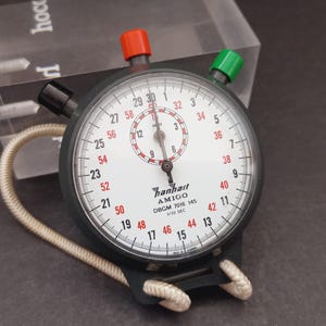 May include: A Hanhart Amigo stopwatch with a white face, black numerals, and red secondary markings. It features a black casing, a red top button, a green side button, and a black side button. A white cord is attached to the base of the device.