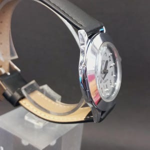 May include: A silver-toned wristwatch with a white face and black hands. The watch has a black leather band and a silver case. The watch is displayed on a clear acrylic stand against a dark background.