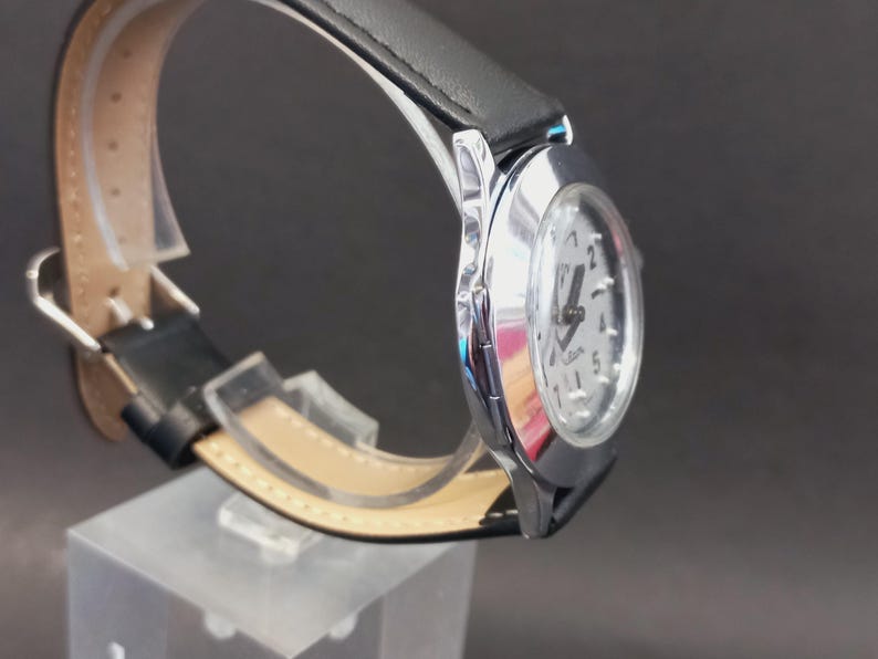 May include: A silver-toned wristwatch with a white face and black hands. The watch has a black leather band and a silver case. The watch is displayed on a clear acrylic stand against a dark background.