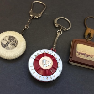 May include: Three vintage keychains. One is a white tire-shaped keychain with a clear center. Another is a red and silver dial-shaped keychain. The third is a brown rectangular keychain with a car image.