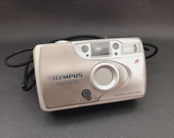 May include: A champagne-colored Olympus Trip AF 50 point-and-shoot film camera with a 28mm lens. The front features the Olympus logo, model name, a built-in flash, an autofocus sensor, and a black wrist strap attached to the side of the body.