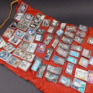 May include: A collection of vintage metal pins with various designs, including portraits and space-themed imagery, displayed on a red fabric. The pins have silver-colored frames and are scattered across the surface.