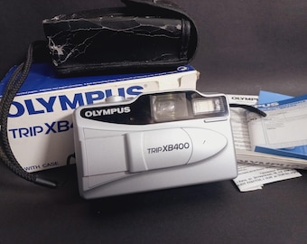 May include: A silver Olympus Trip XB400 camera with a black leather case and box. The camera has a built-in flash and a lens. The box is blue and white with the Olympus logo. Includes a black camera strap and instruction manuals.