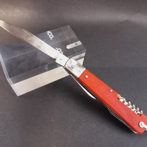 Op de afbeelding: Een inklapbaar zakmes met een uitgeklapt zilverkleurig metalen lemmet en een felrood handvat. Het handvat is voorzien van een metalen bolster met een spiraalvormige gravure, een zilveren kurkentrekker en een kleine metalen ring aan de basis.